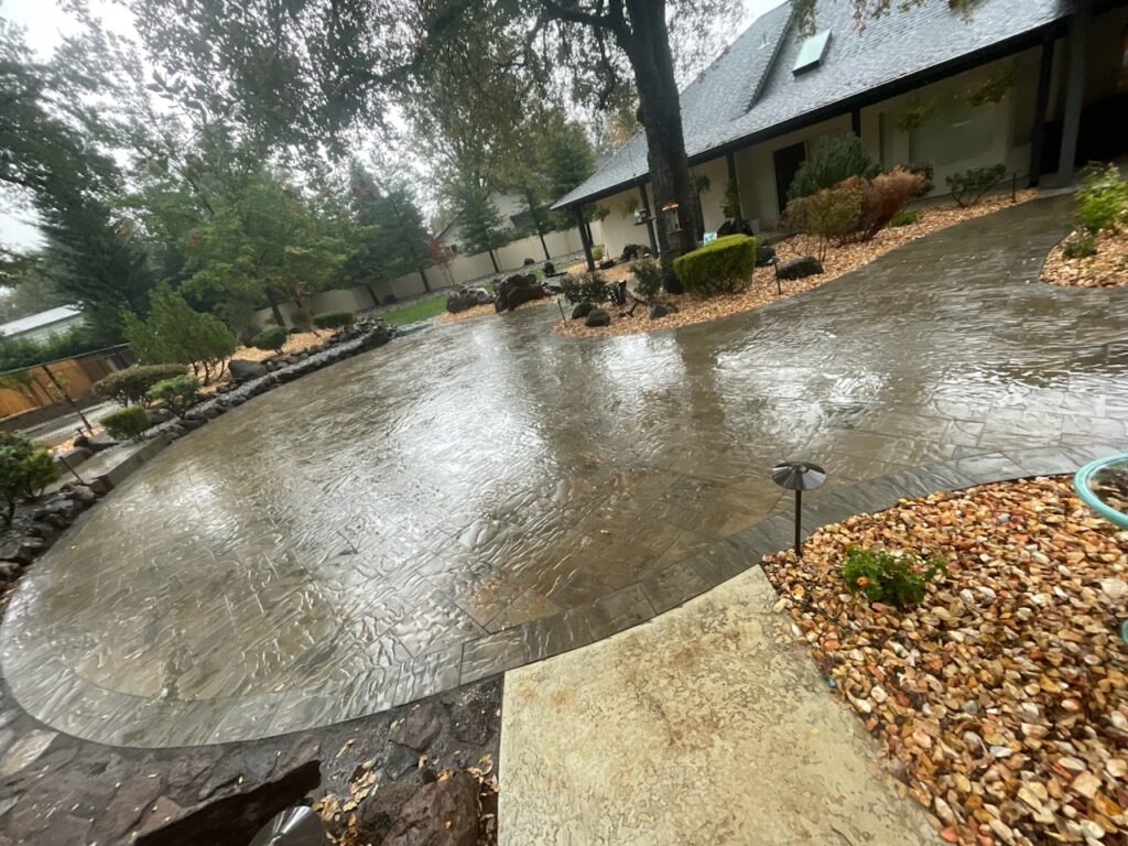 Wide stone patio area surrounded by landscaped garden with trees and shrubs beside a house exterior on a rainy day creating a peaceful and serene outdoor setting - work by Mijangos Professional Landscape Services LLC in Redding, CA