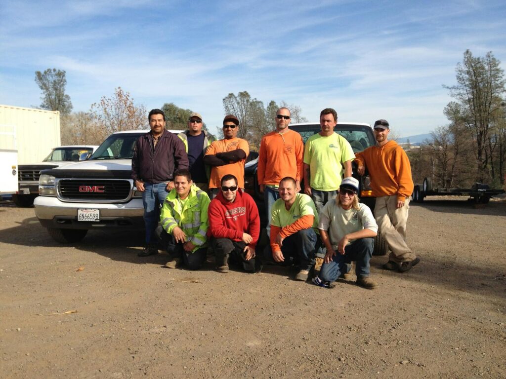 A group of workers in casual clothes standing and kneeling in front of two trucks outdoors on a sunny day with trees and a clear blue sky in the background. - work by Mc Entire Landscaping in Redding, CA