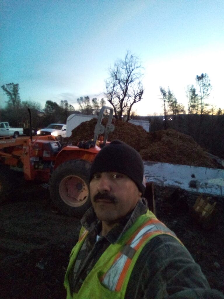 Worker in safety vest and beanie takes a self-portrait at a worksite during dawn standing in front of an orange tractor with trees and a clear sky in the background - work by Mc Entire Landscaping in Redding, CA