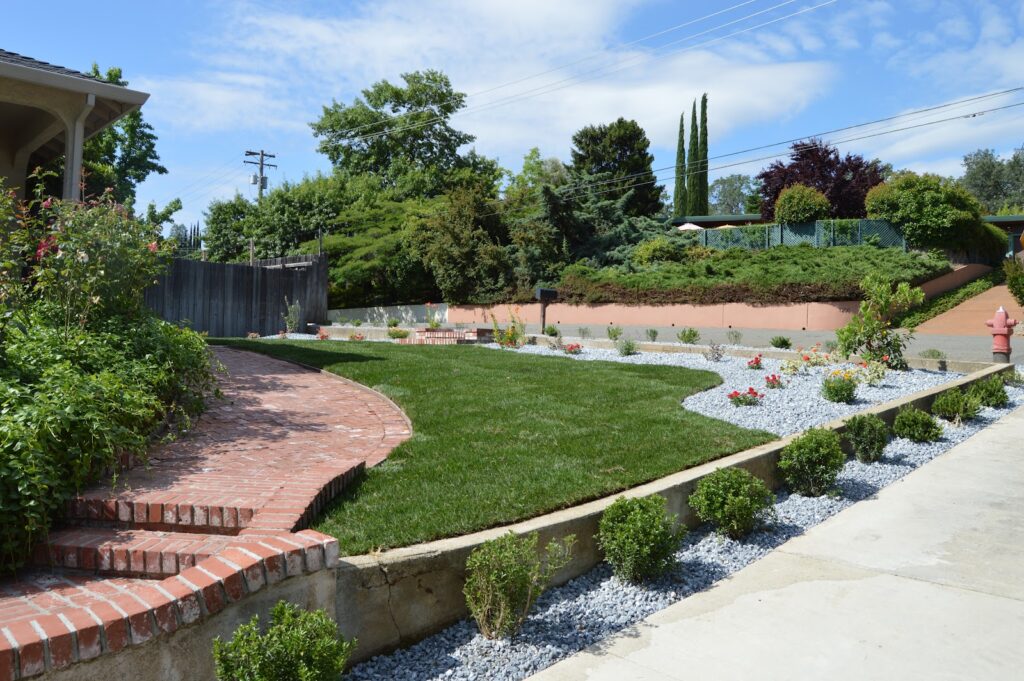 Beautiful landscaped backyard with green grass, decorative shrubs, stone pathway, and brick steps under a clear blue sky, showcasing a well-maintained garden design. - work by Clifford Landscape Contractors in Redding, CA
